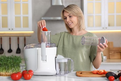 Smiling woman putting fresh tomato into juicer at white marble table in kitchen