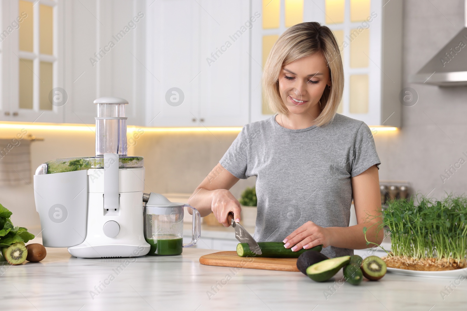 Photo of Juicer and fresh products on white marble table. Smiling woman cutting cucumber in kitchen