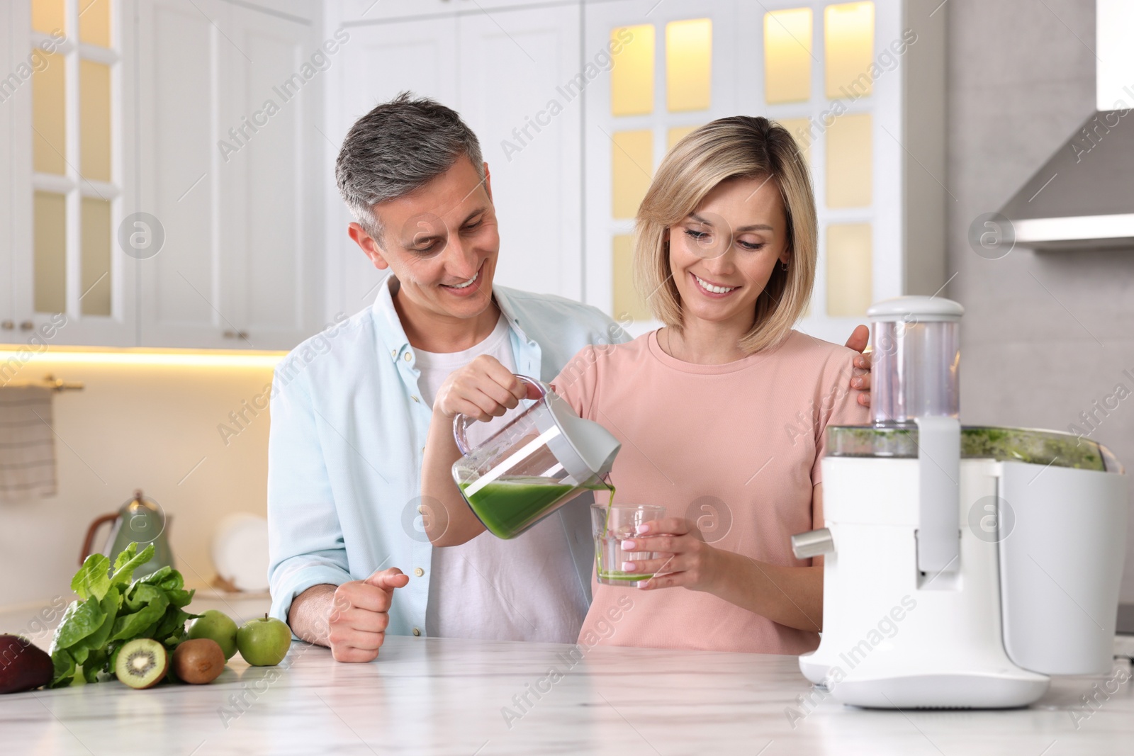 Photo of Happy couple with juicer, fresh products and tasty juice at white marble table in kitchen