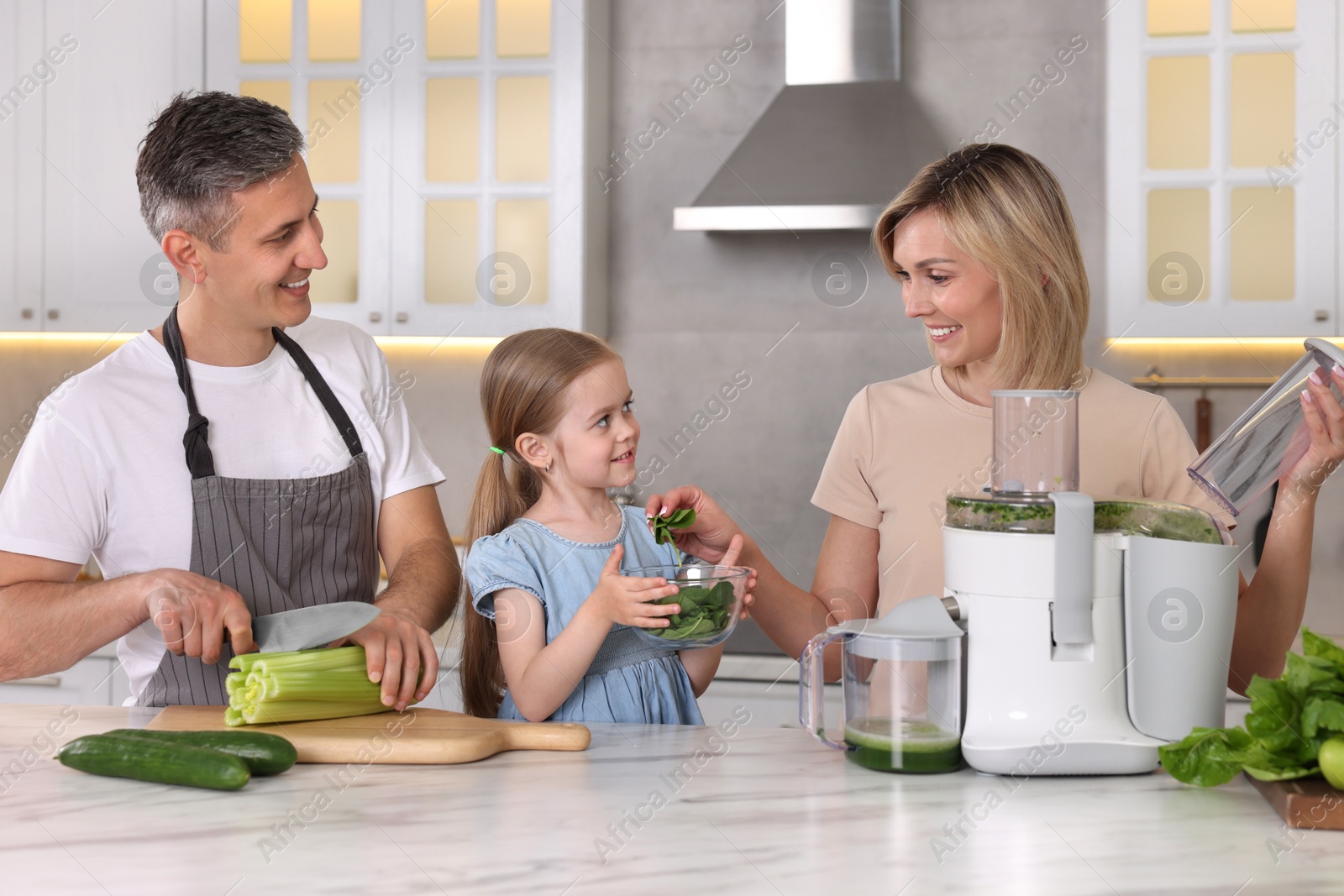 Photo of Happy family with juicer and fresh products making drink at white marble table in kitchen