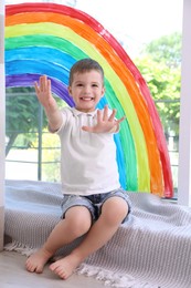 Photo of Little boy near rainbow painting on window indoors