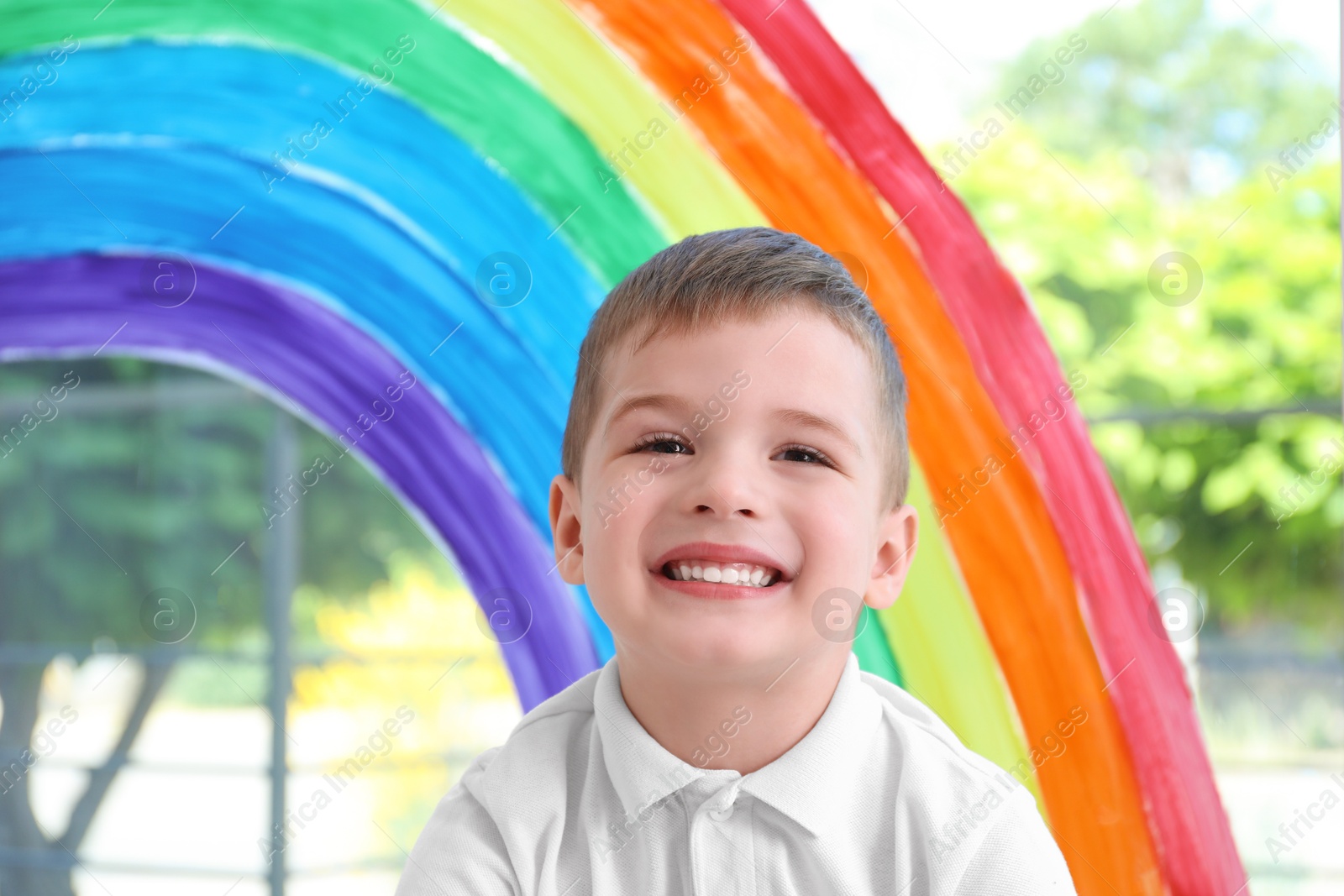 Photo of Little boy near rainbow painting on window indoors