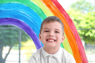 Photo of Little boy near rainbow painting on window indoors