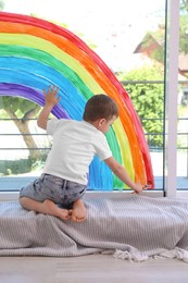 Photo of Little boy drawing rainbow on window indoors