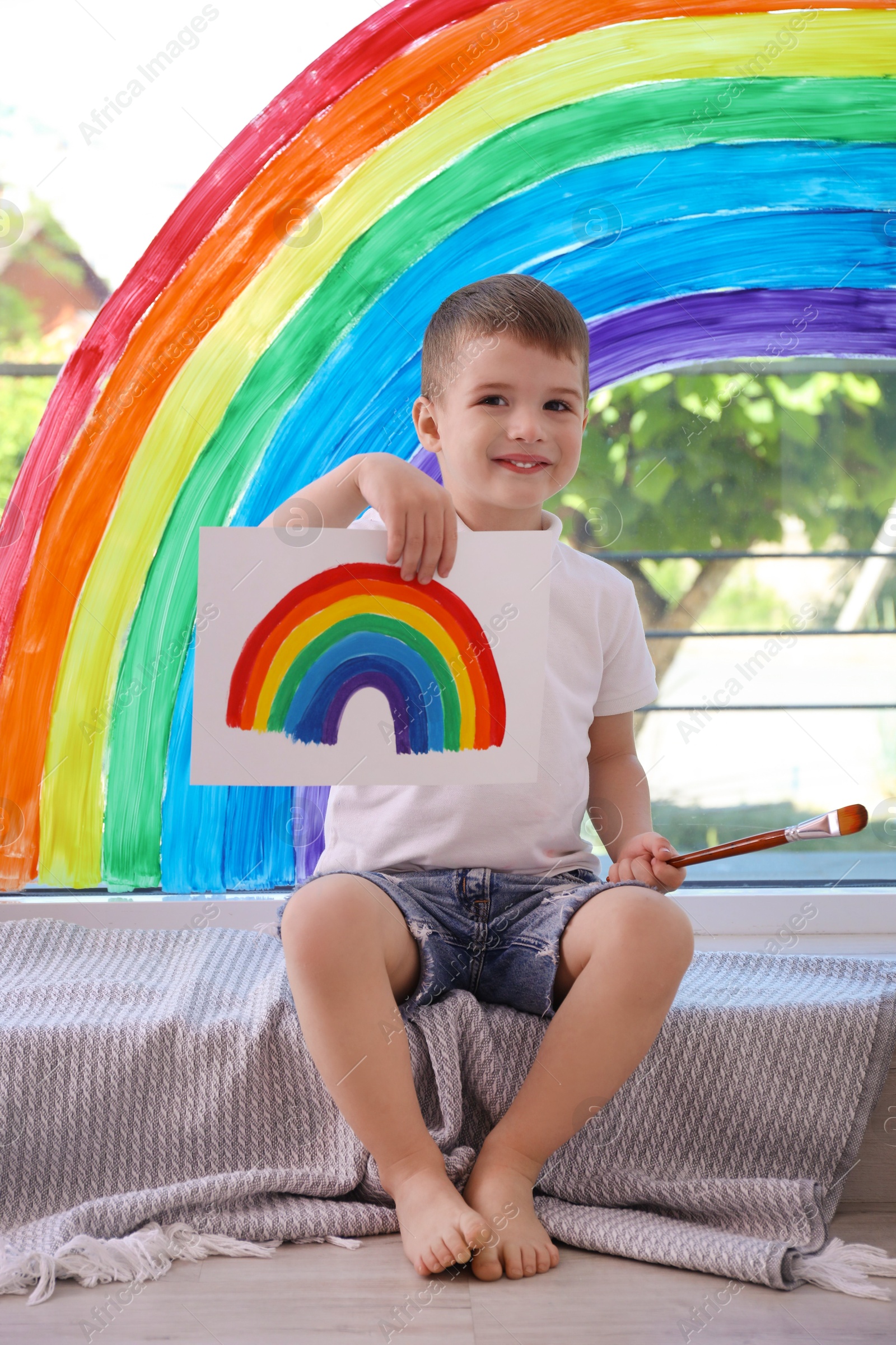 Photo of Happy little boy with picture of rainbow and brush near window indoors