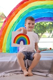 Photo of Happy little boy with picture of rainbow and brush near window indoors