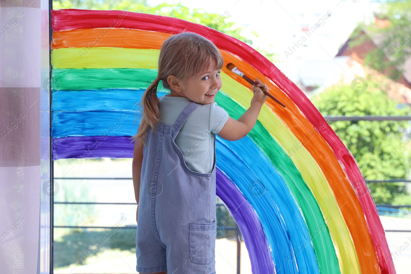 Photo of Little girl drawing rainbow on window indoors