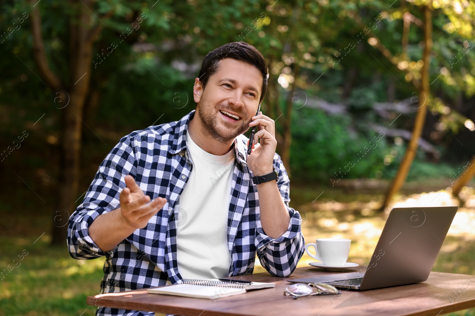 Photo of Smiling freelancer talking on smartphone at table with laptop outdoors. Remote job