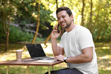 Smiling freelancer talking on smartphone at table with laptop outdoors. Remote job