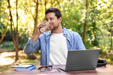 Smiling freelancer drinking coffee at table with laptop outdoors. Remote job