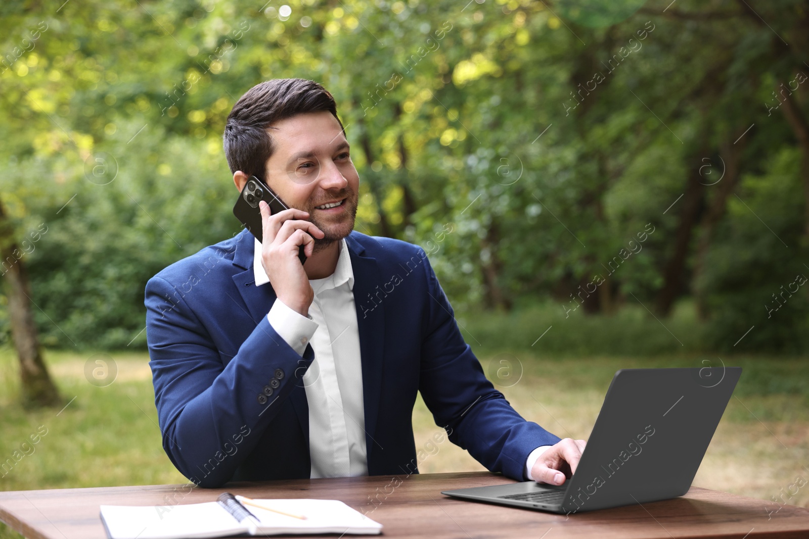 Photo of Smiling businessman talking on smartphone at table with laptop outdoors. Remote job