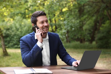 Smiling businessman talking on smartphone at table with laptop outdoors. Remote job