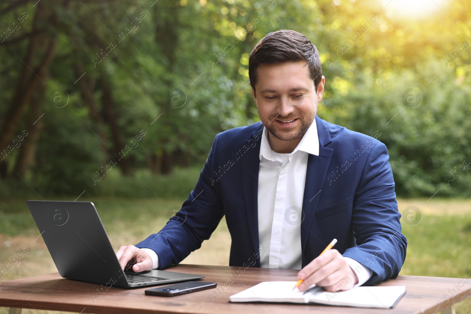 Photo of Smiling businessman working with laptop and writing something at table outdoors. Remote job