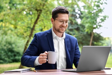 Photo of Smiling businessman with cup of coffee working with laptop at table outdoors. Remote job