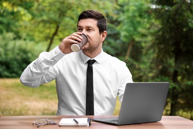 Photo of Businessman drinking coffee at table with laptop outdoors. Remote job