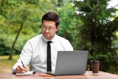 Photo of Businessman working with laptop and writing something at table outdoors. Remote job