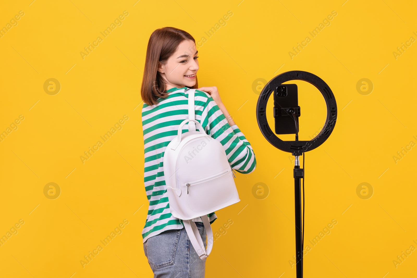 Photo of Fashion blogger reviewing backpack and recording video with smartphone and ring lamp on orange background