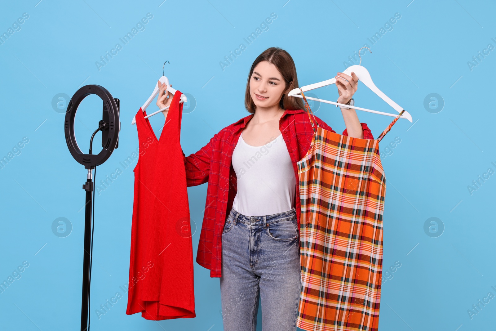 Photo of Fashion blogger reviewing dresses and recording video with smartphone and ring lamp on light blue background