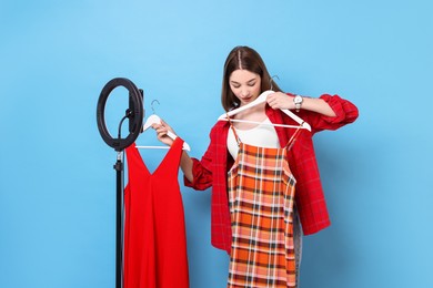Photo of Fashion blogger reviewing dresses and recording video with smartphone and ring lamp on light blue background