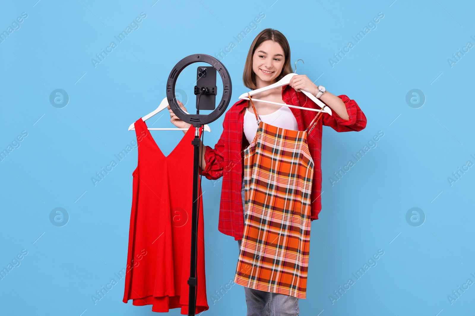 Photo of Fashion blogger reviewing dresses and recording video with smartphone and ring lamp on light blue background