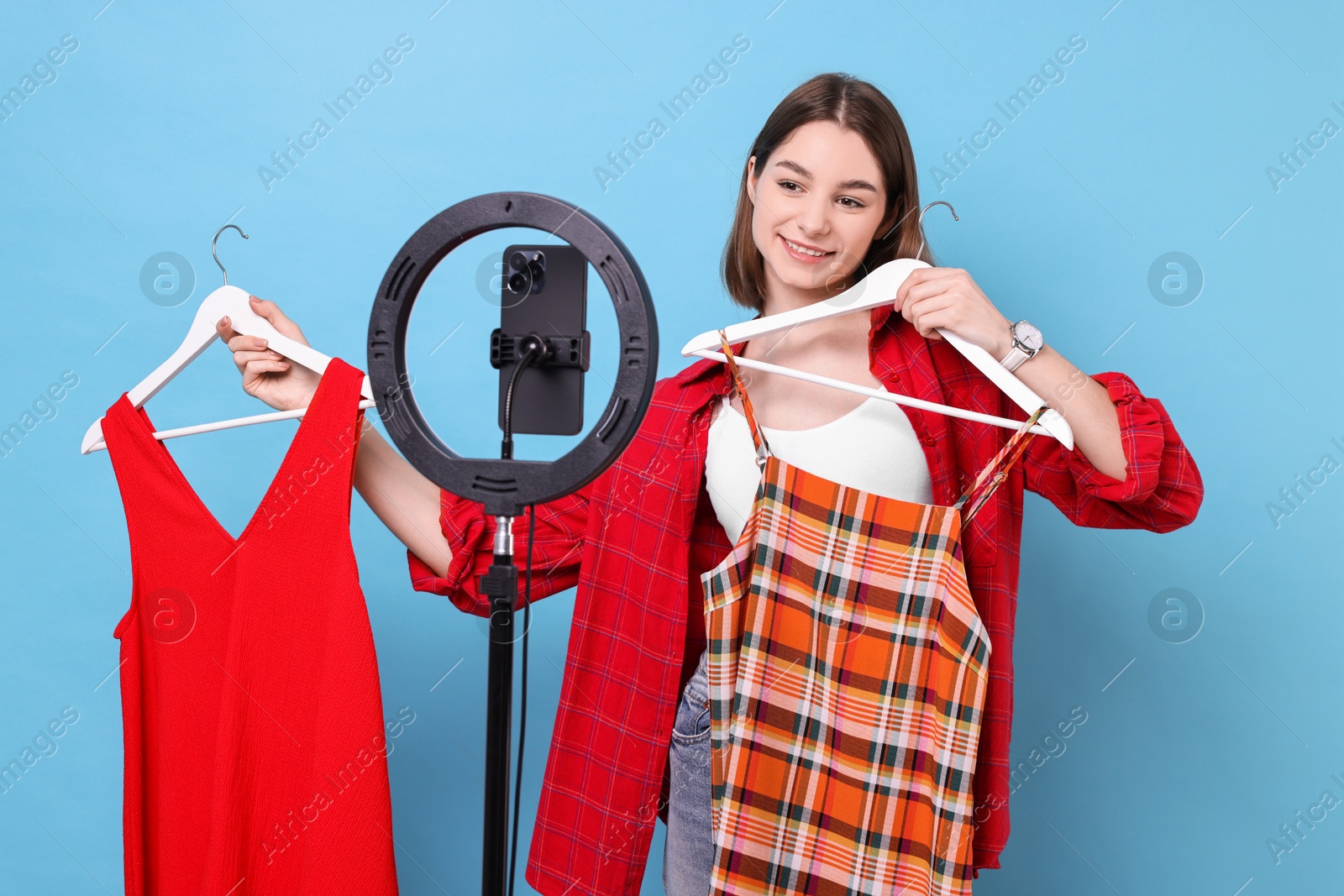 Photo of Fashion blogger reviewing dresses and recording video with smartphone and ring lamp on light blue background