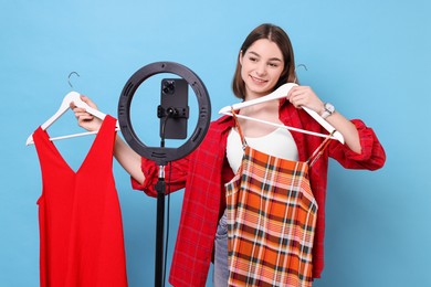 Photo of Fashion blogger reviewing dresses and recording video with smartphone and ring lamp on light blue background