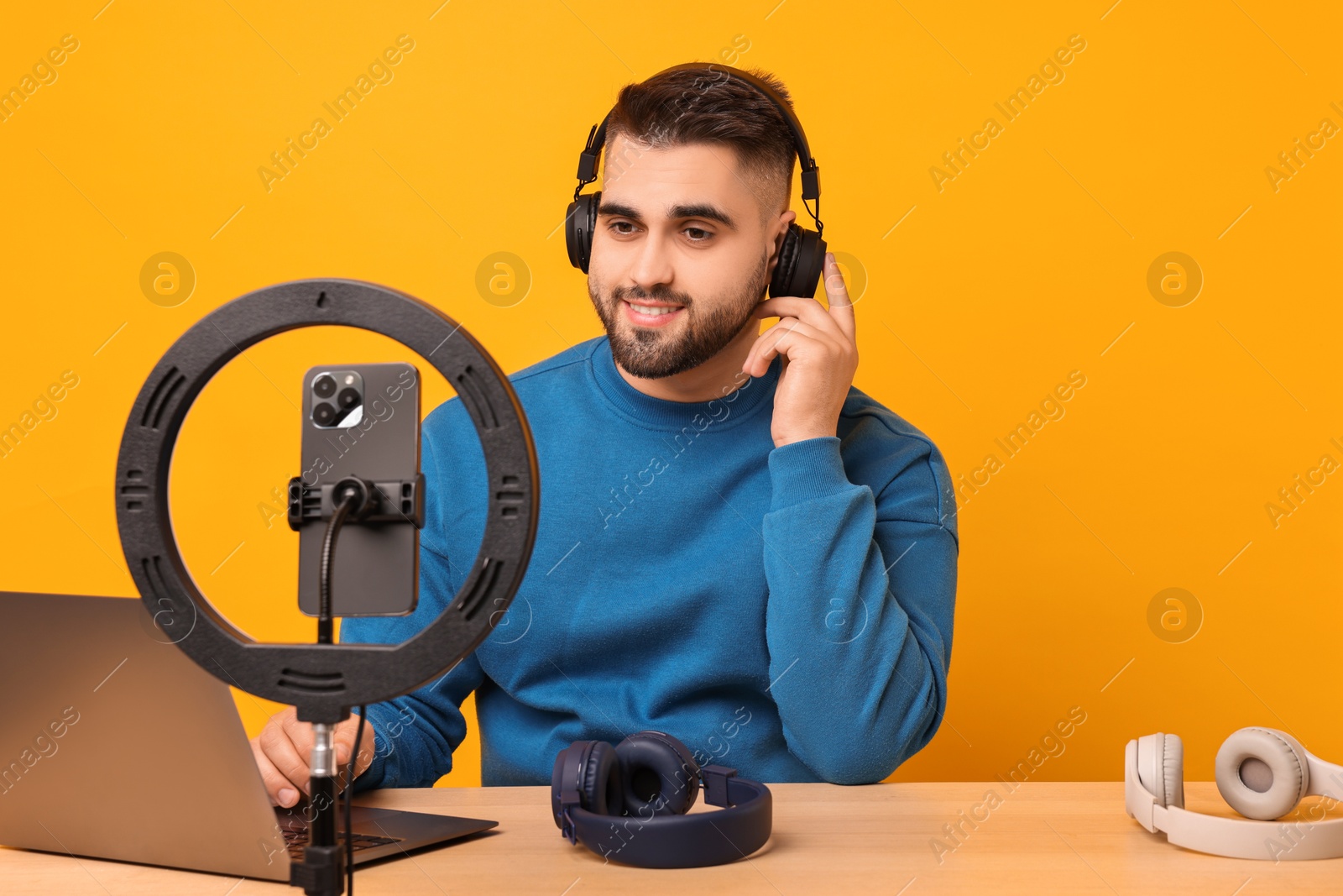 Photo of Technology blogger reviewing headphones and recording video with smartphone and ring lamp at wooden table on orange background