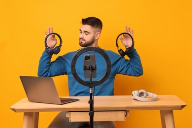 Photo of Technology blogger reviewing headphones and recording video with smartphone and ring lamp at wooden table on orange background