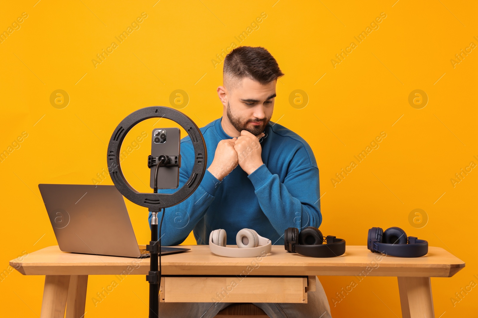 Photo of Technology blogger reviewing headphones and recording video with smartphone and ring lamp at wooden table on orange background