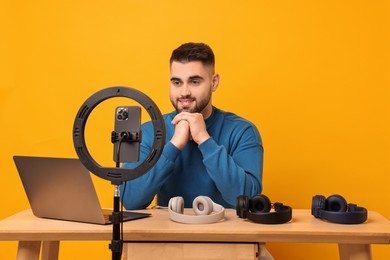 Photo of Technology blogger reviewing headphones and recording video with smartphone and ring lamp at wooden table on orange background