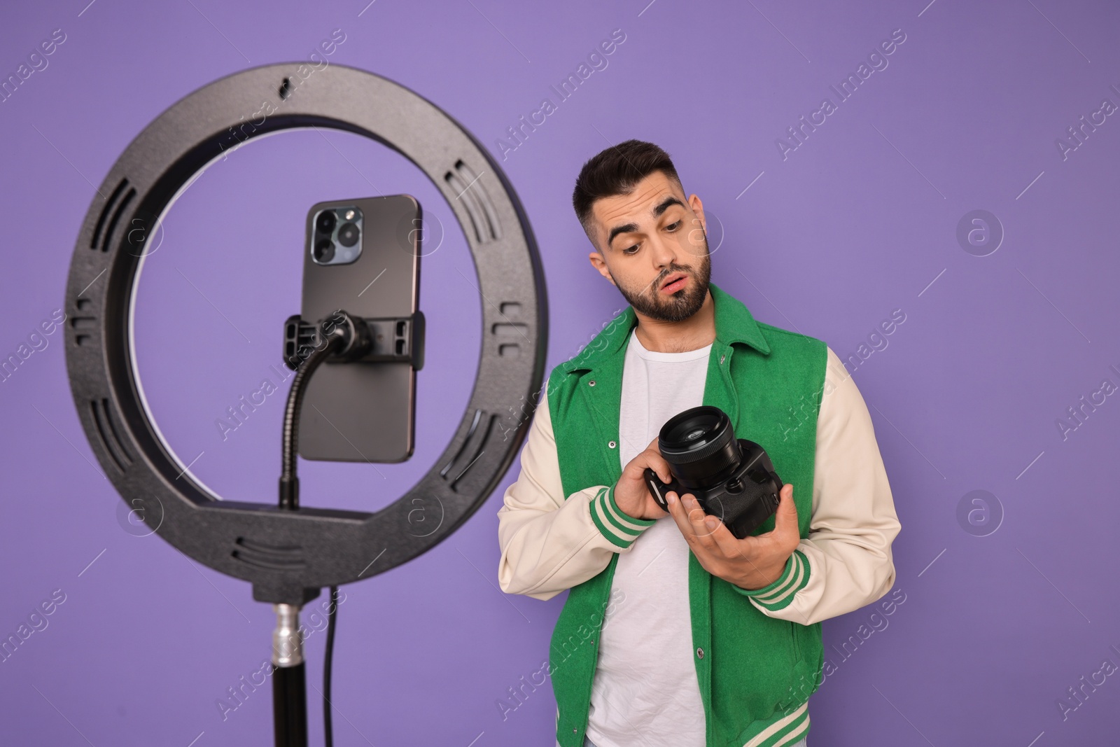 Photo of Technology blogger reviewing camera and recording video with smartphone and ring lamp on purple background