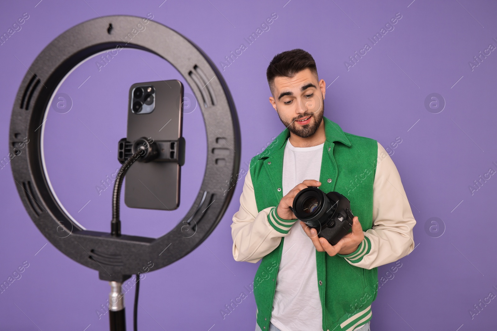 Photo of Technology blogger reviewing camera and recording video with smartphone and ring lamp on purple background