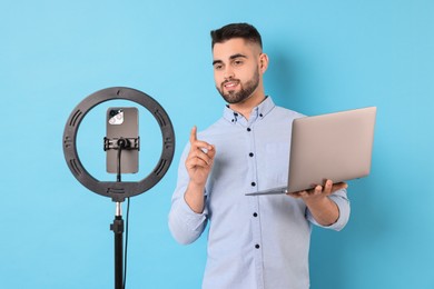 Photo of Technology blogger reviewing laptop and recording video with smartphone and ring lamp on light blue background
