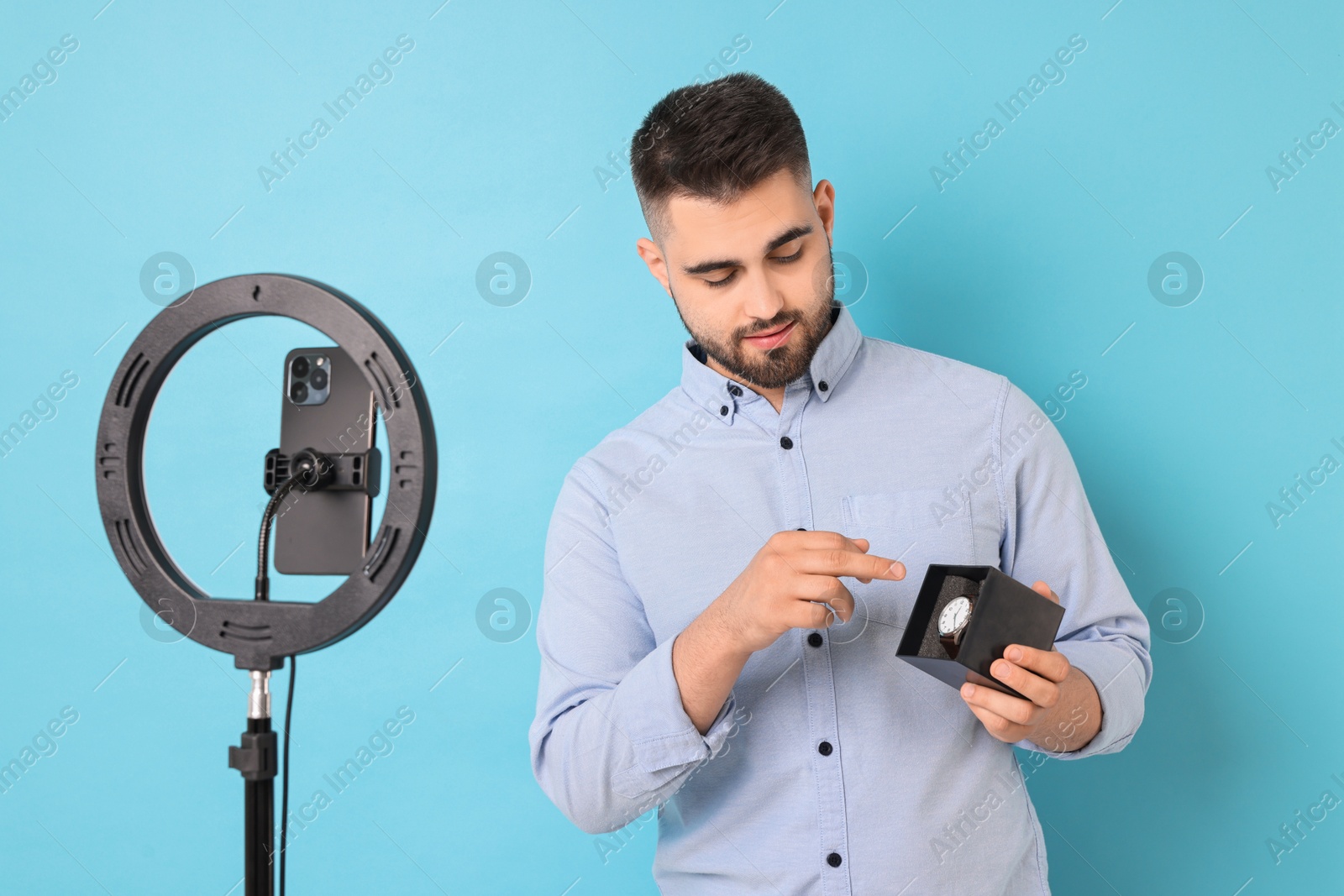 Photo of Fashion blogger reviewing watch and recording video with smartphone and ring lamp on light blue background