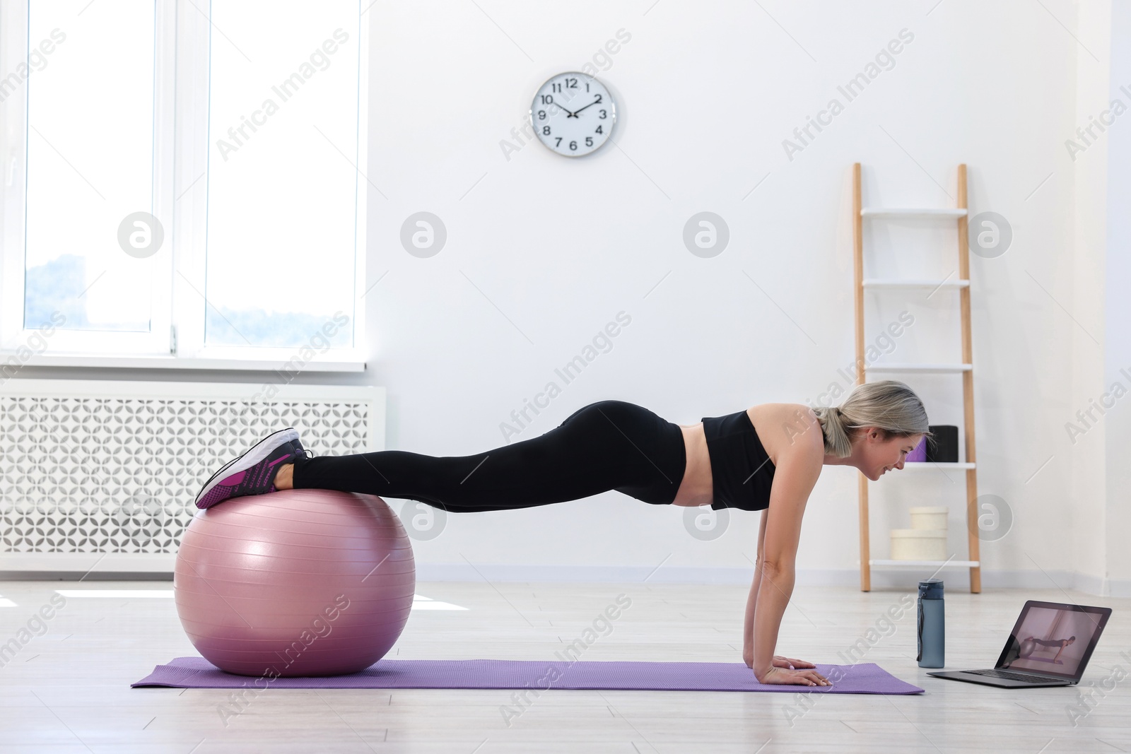 Photo of Online fitness trainer. Woman doing exercise with fitness ball near laptop at home