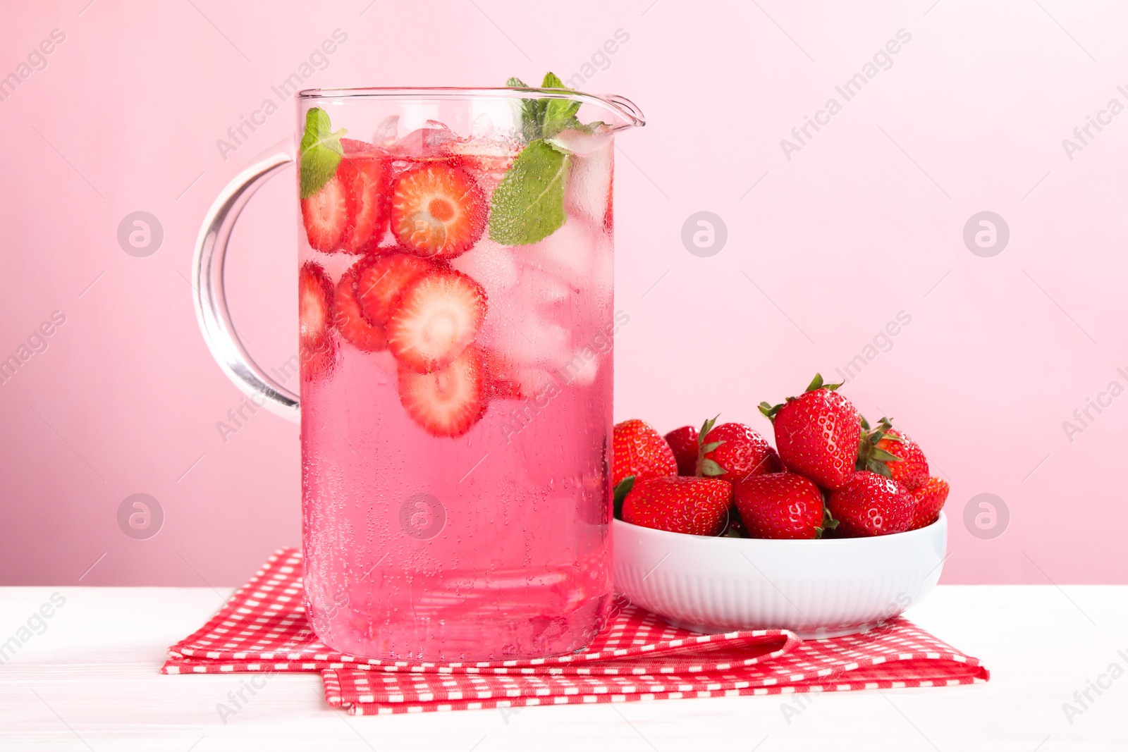 Photo of Freshly made strawberry lemonade with mint in jug on white wooden table against pink background