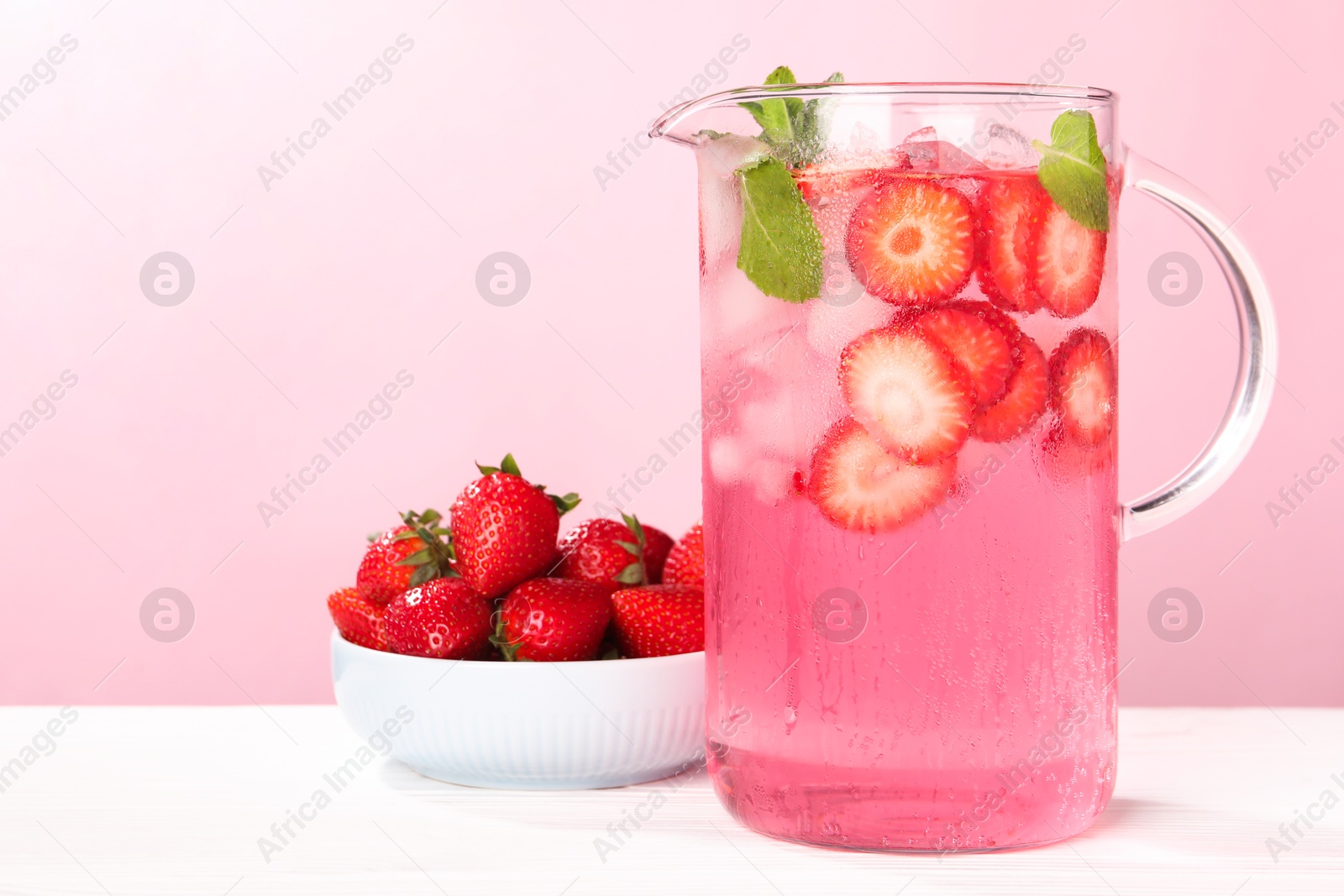 Photo of Freshly made strawberry lemonade with mint in jug on white wooden table against pink background