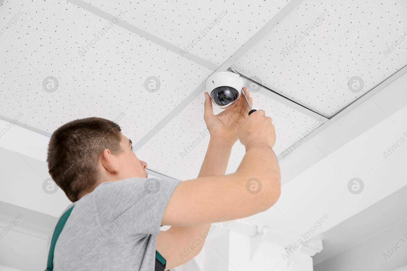 Photo of Technician with screwdriver installing CCTV camera on ceiling indoors, low angle view