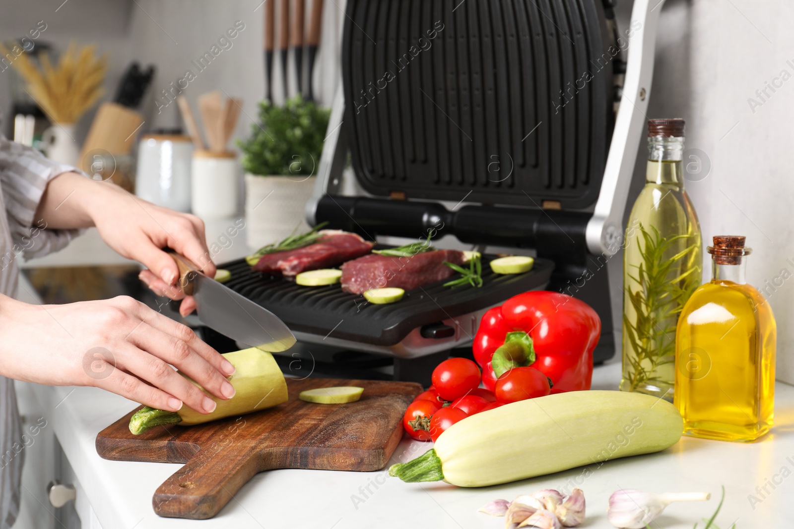 Photo of Woman cooking different products with electric grill at white wooden table in kitchen, closeup