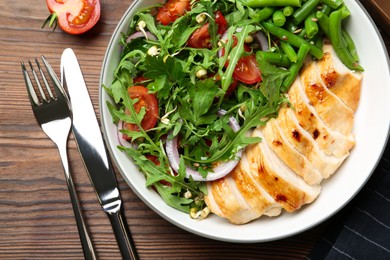 Photo of Healthy meal. Tasty salad, cutlery and chicken breast in bowl on wooden table, flat lay