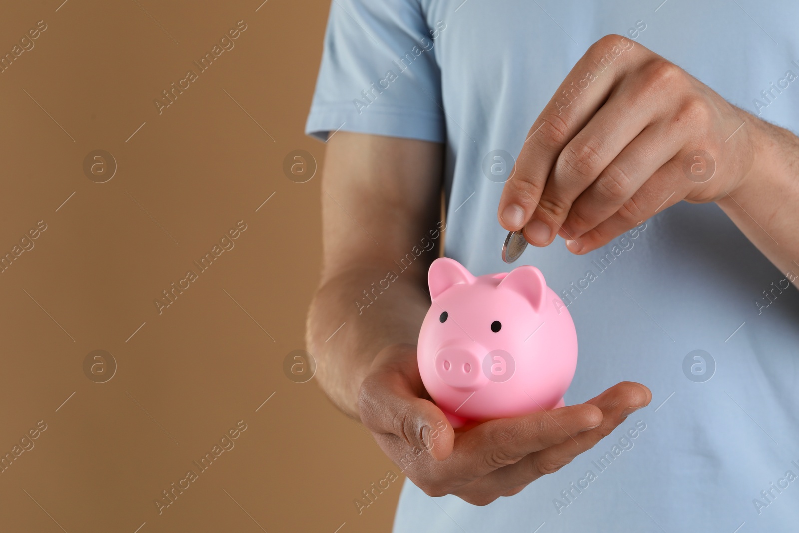 Photo of Man putting coin into pink piggy bank on light brown background, closeup. Space for text