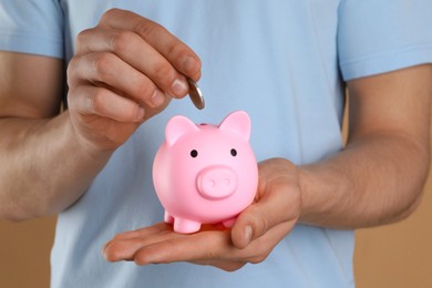 Photo of Man putting coin into pink piggy bank on light brown background, closeup