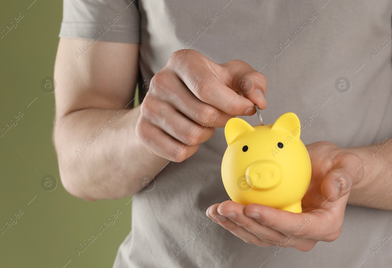 Photo of Man putting coin into yellow piggy bank on olive background, closeup