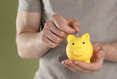 Man putting coin into yellow piggy bank on olive background, closeup