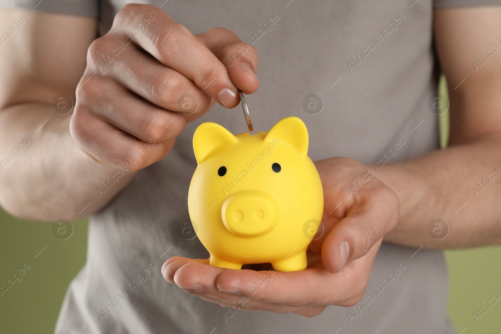 Photo of Man putting coin into yellow piggy bank on olive background, closeup