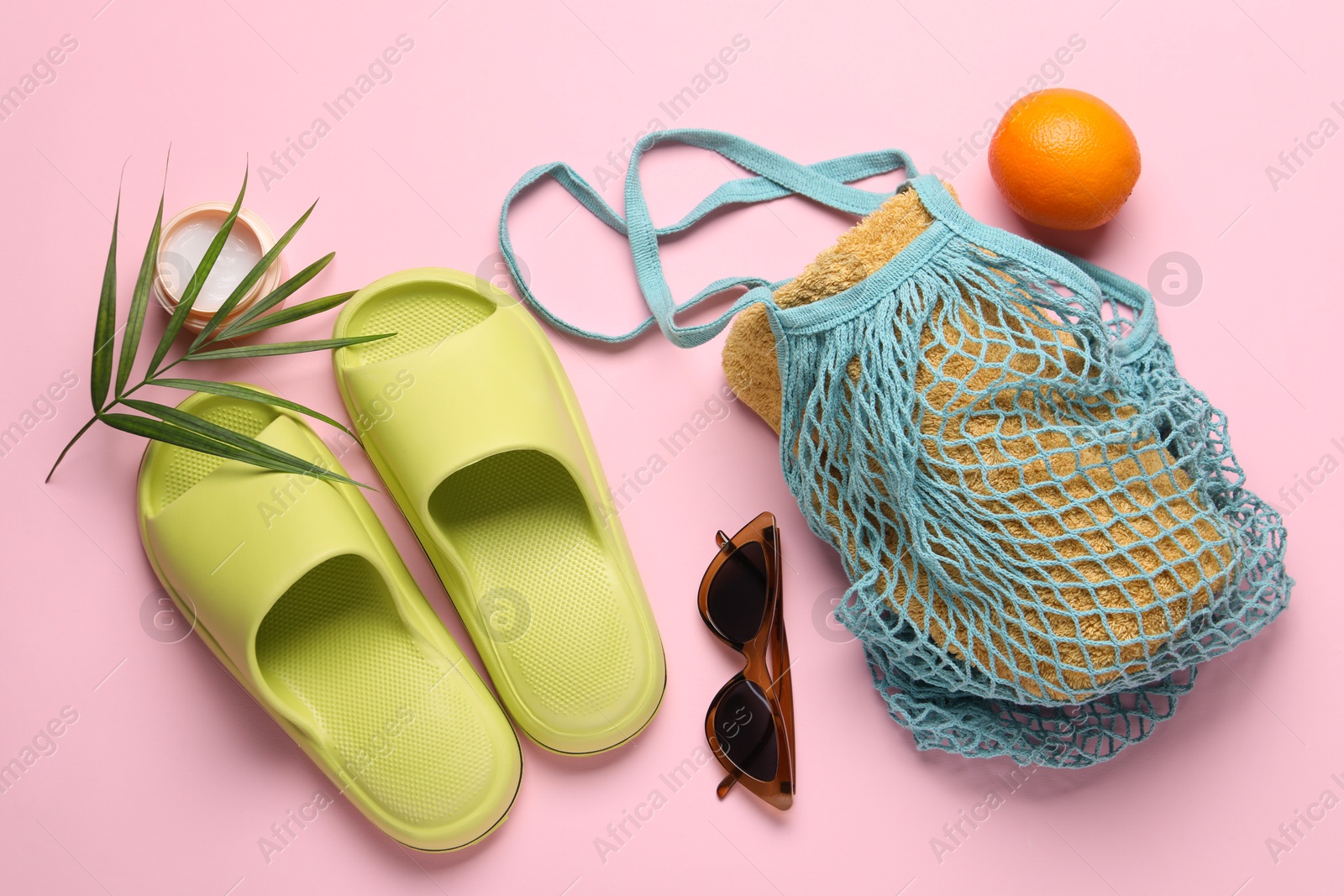 Photo of String bag with towel, sunglasses, orange, slippers, cream and palm leaf on pink background, flat lay