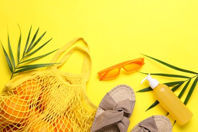 String bag with oranges, sunglasses, cosmetic product, slippers and palm leaves on yellow background, flat lay