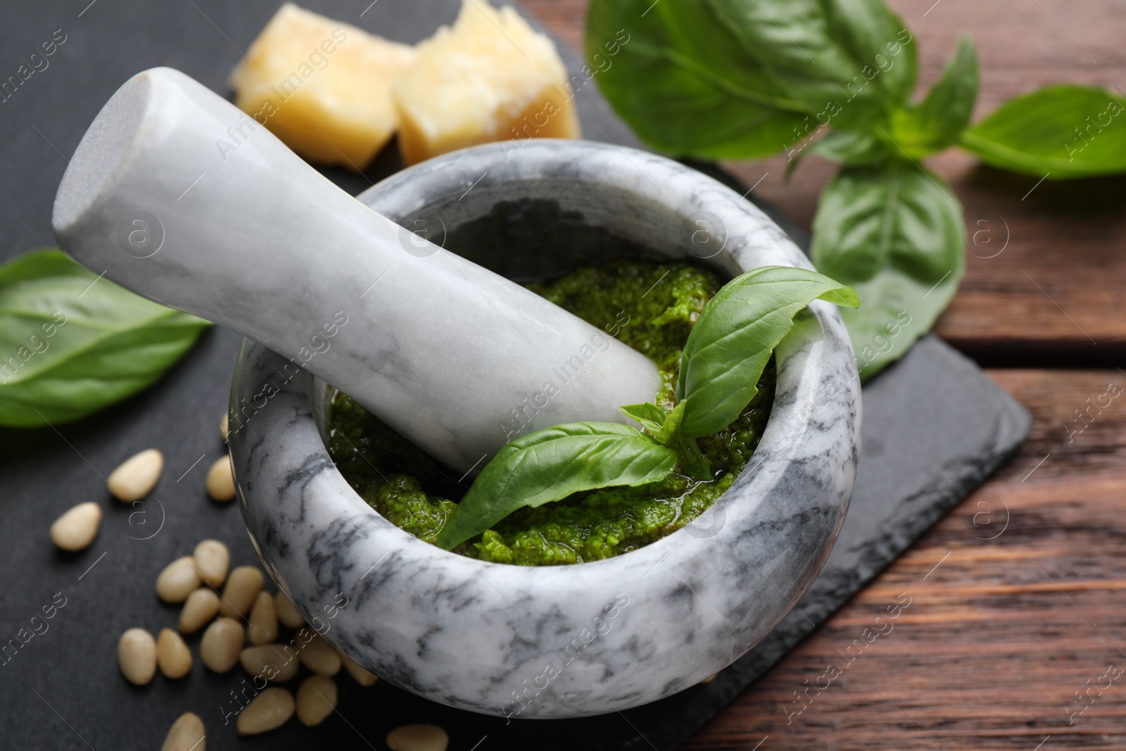 Photo of Tasty pesto sauce in mortar, pestle, basil, pine nuts and cheese on wooden table, closeup