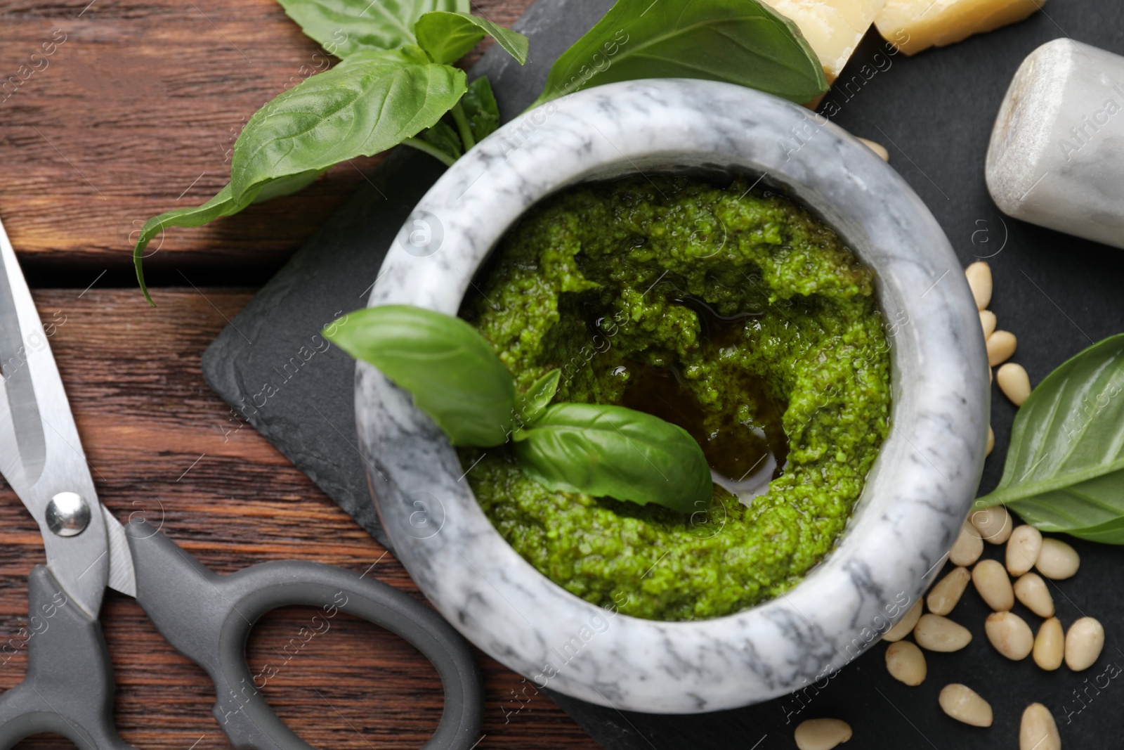 Photo of Tasty pesto sauce in mortar, basil, pine nuts and scissors on wooden table, top view