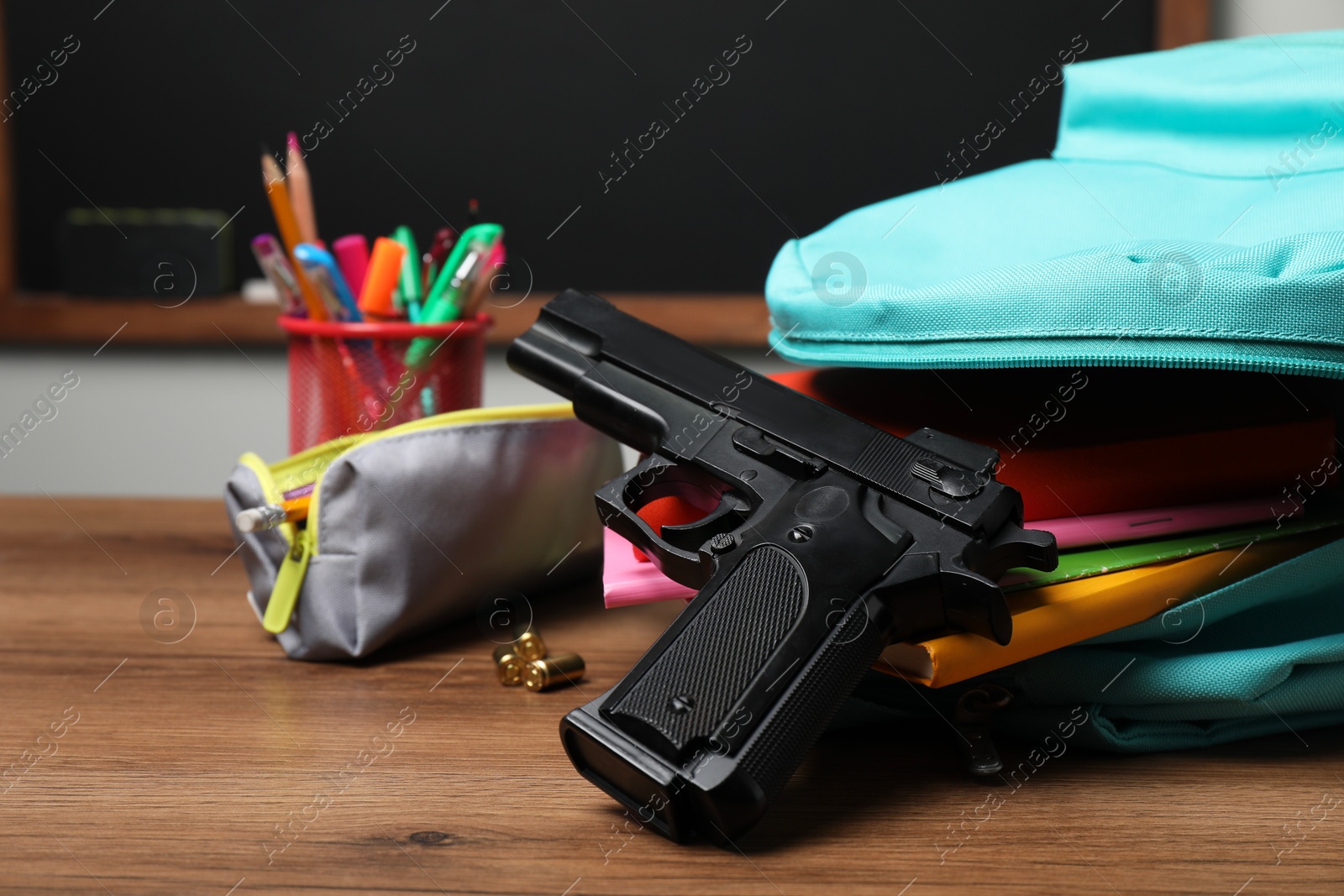 Photo of Gun, bullets and school stationery on wooden table near blackboard indoors, closeup